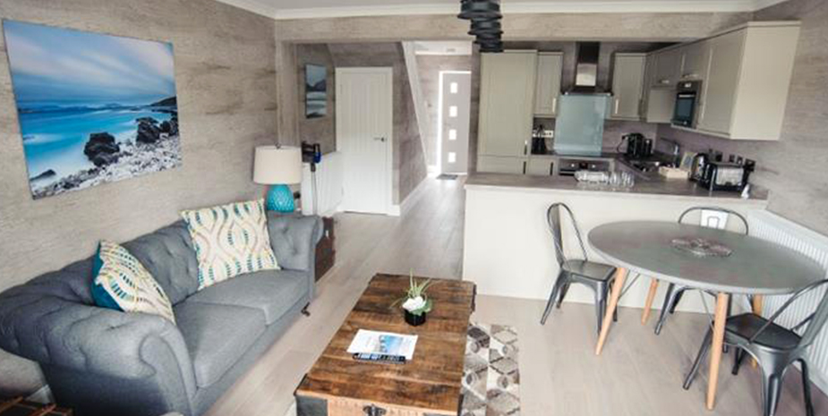 Kirklea Island Suites Lounge and kitchen with grey sofa, seating area and kitchen at rear.