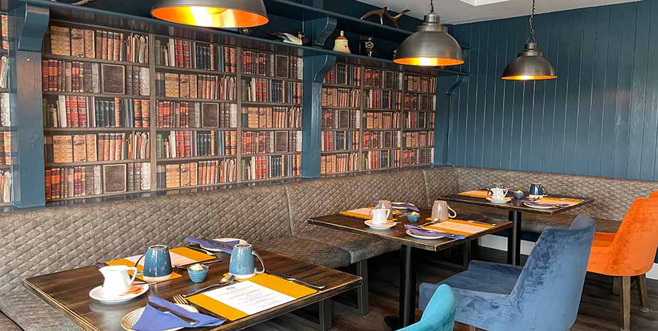 Grianaig Dining Room with bench style seating and atables set uwith contrast colour chairs, teal wall panelling and bookcase style wallpaper.