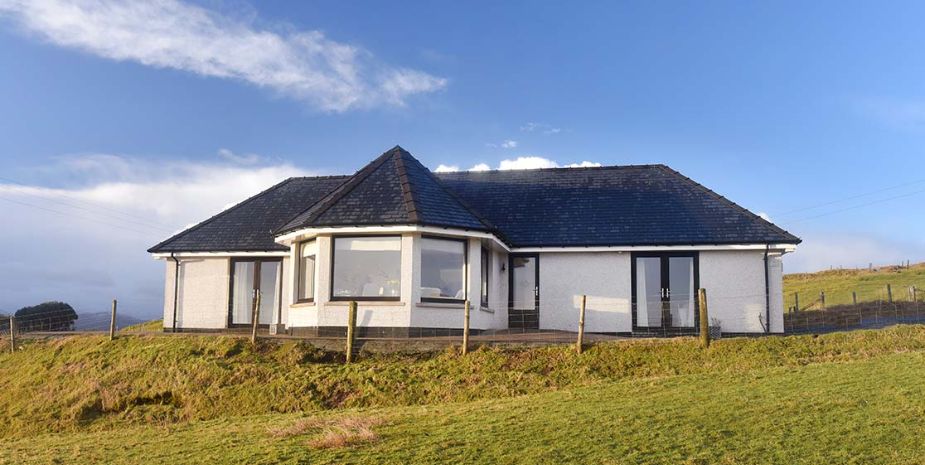 Taigh Chailean single-story white house with dark tiled roof and large windows, located in a rural area with a grassy field and a clear blue sky overhead.