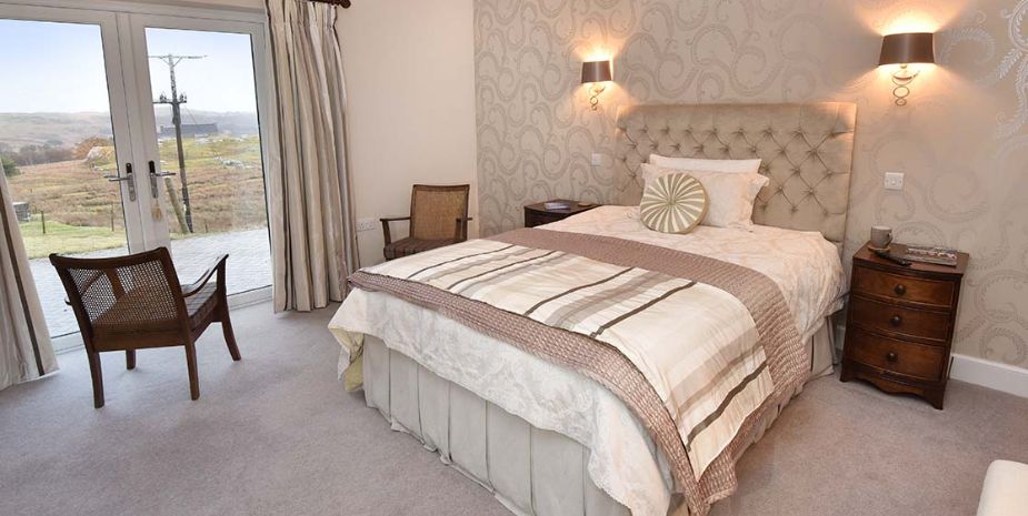 A neatly arranged bedroom featuring an upholstered headboard, patterned wallpaper, two wall mounted lights, and a coordinated bedding set with decorative pillows.