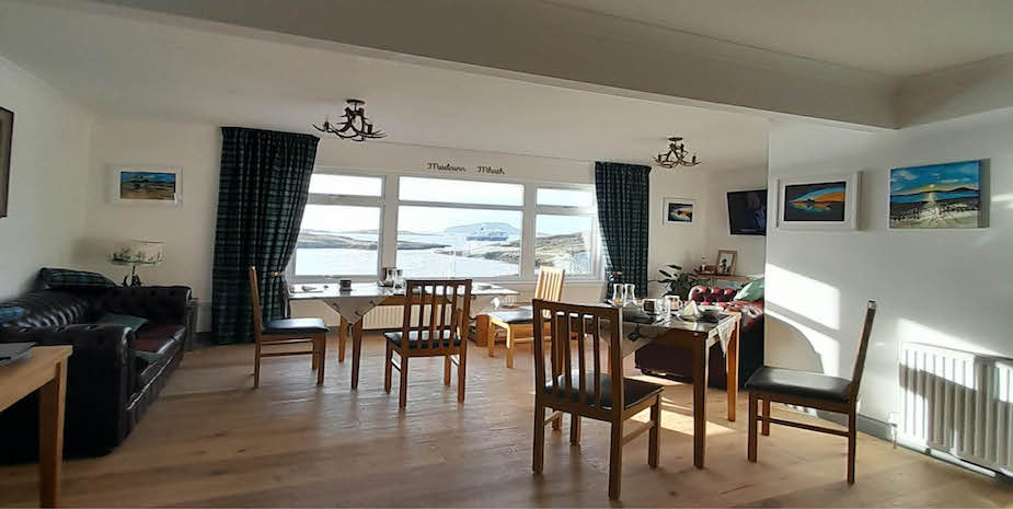 Bright dining space at Bayview Guest House with tables and chairs arranged near wide windows overlooking a Castlebay. Artwork of coastal scenes hangs on the white walls, and plaid curtains add a touch of warmth to the room, which has a mix of traditional and rustic decor.