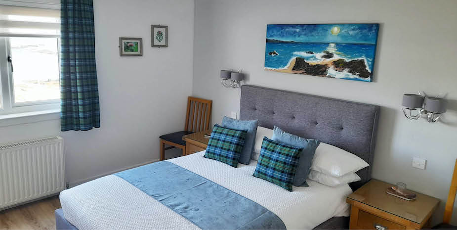 Well lit bedroom in Bayview Guest House featuring a grey headboard with check pillows and a soft blue bed runner. Vibrant coastal artwork hangs above the headboard, and the room is finished with wood bedside tables and matching check curtains that frame a sunlit window.