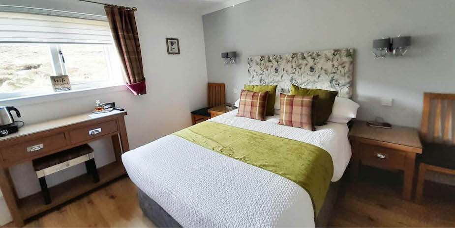 Warm toned bedroom in Bayview Guest House featuring a floral headboard and a double bed with check and green accent pillows. Check curtains complements the decor, and there’s a small wooden desk under the window.