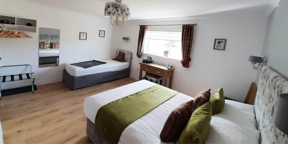 Warm toned bedroom in Bayview Guest House featuring a floral headboard and a double bed wit single bed with check and green accent pillows. Check curtains complements the decor, and there’s a small wooden desk under the window.